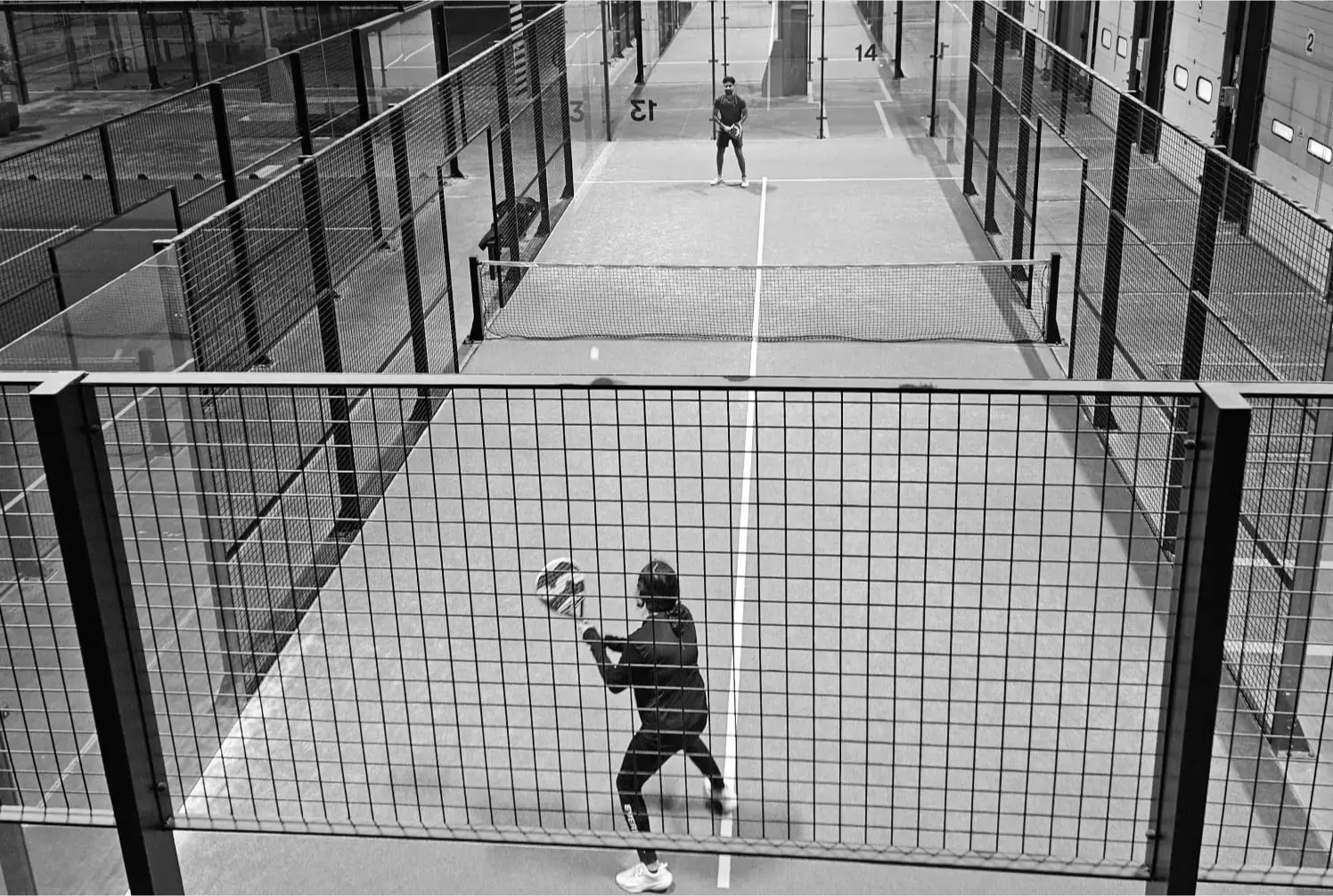 Black-and-white photo of two players on a single indoor padel court, separated by netting and surrounded by high fences.