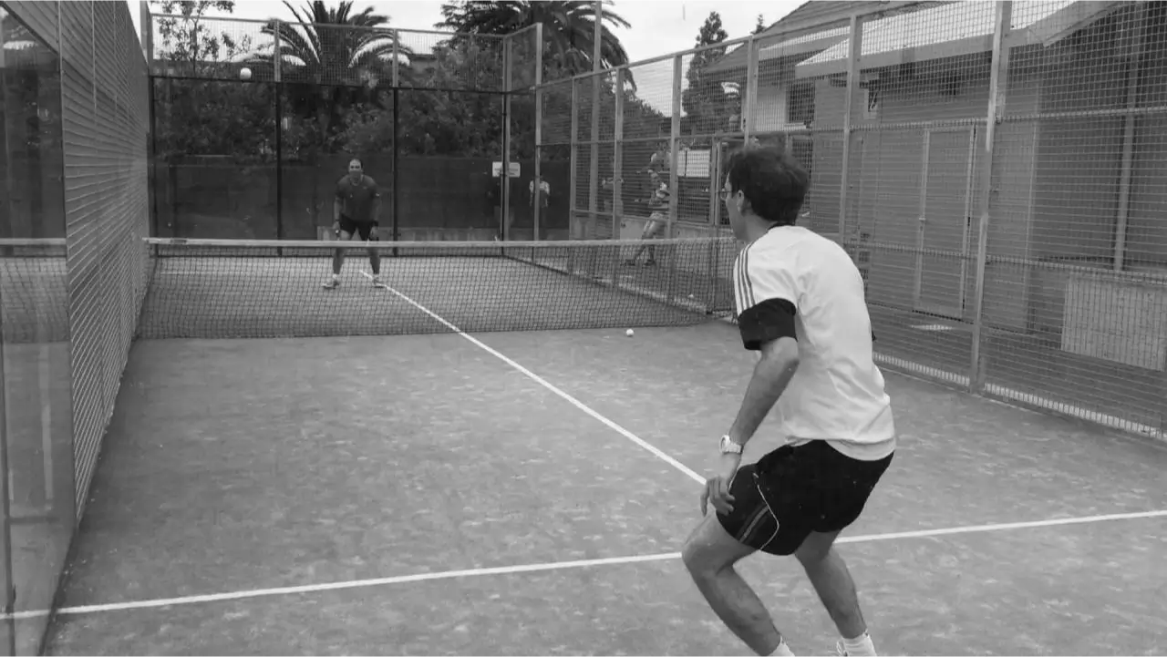 Two players on a single outdoor padel court mid-game, surrounded by fencing and trees in the background.