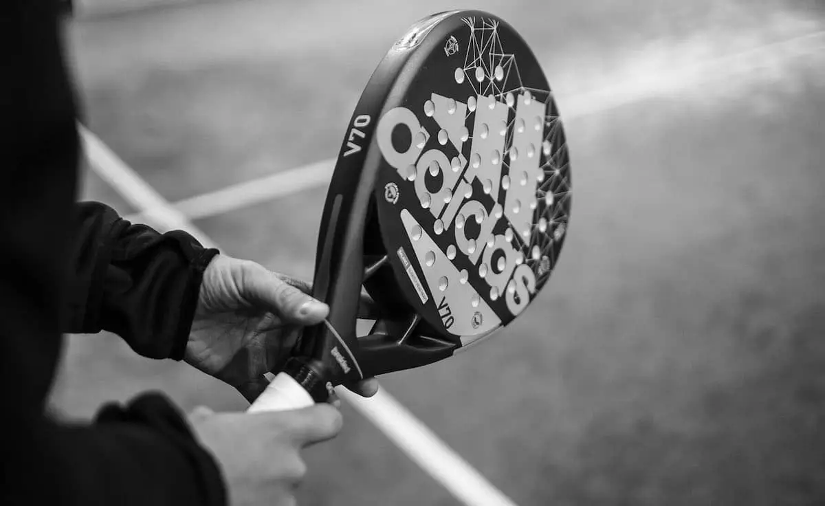 A person gripping an Adidas padel racket with both hands on a padel court, preparing for a shot.