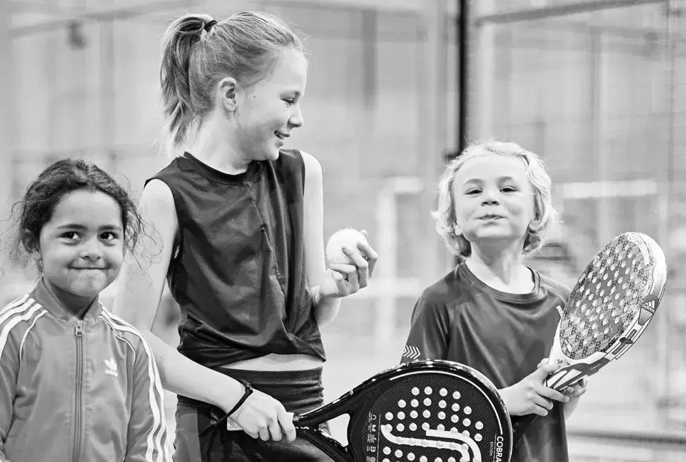 Three smiling kids holding padel rackets and a ball, enjoying a fun padel session together on the court.