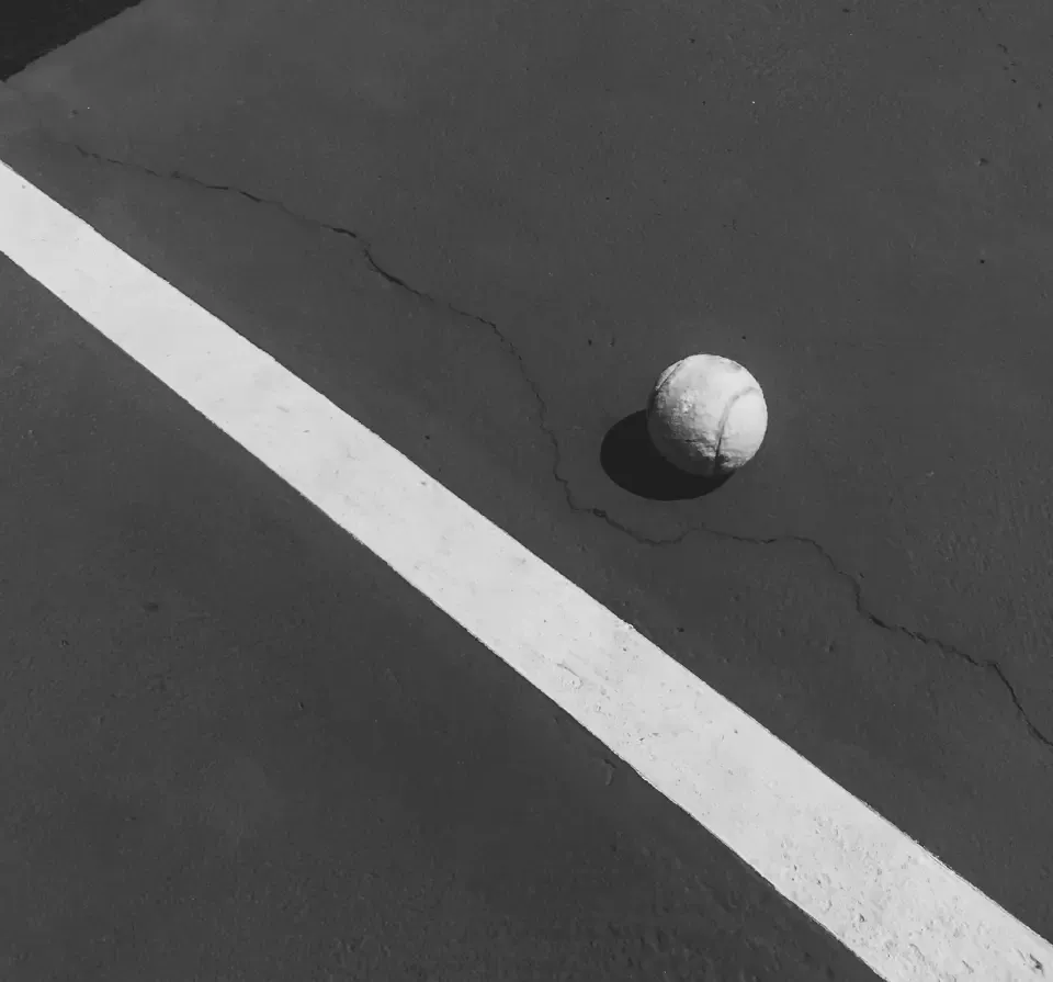 Black and white image of a tennis ball on a cracked concrete court with a bold white line.