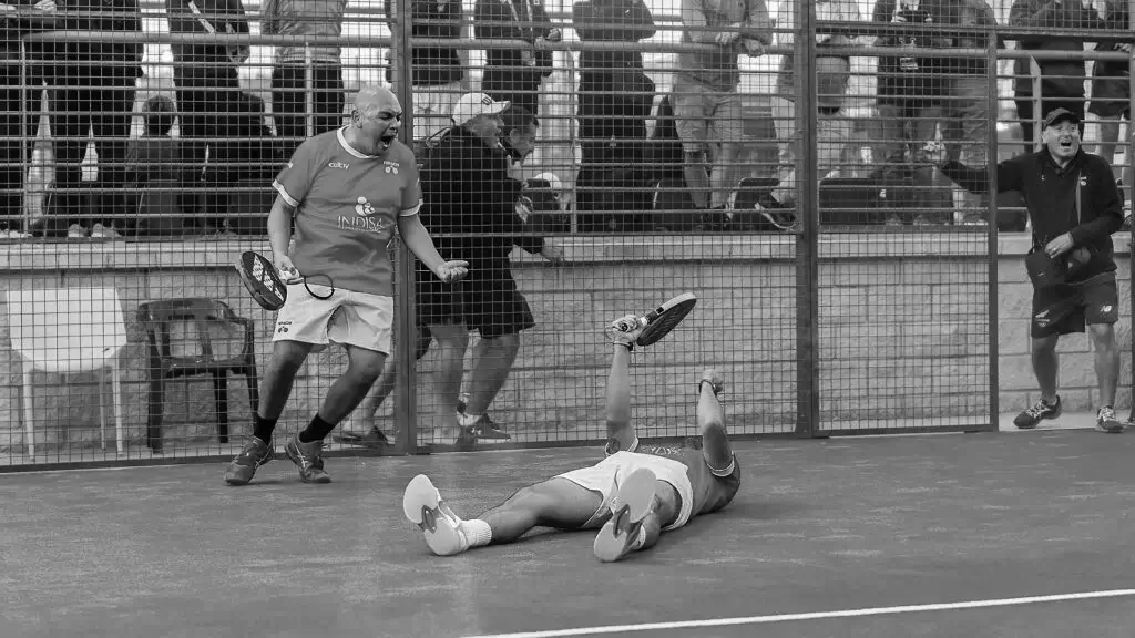 Two padel players celebrate an intense victory, with one lying on the court and the other cheering passionately near the fence.